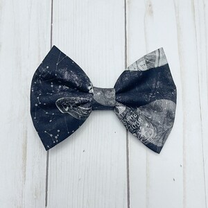 May include: A fabric bow tie featuring a dark navy blue background with a white celestial print of constellations, stars, and planetary or lunar imagery. The bow tie has a classic pleated center knot and pointed wing edges on both sides.