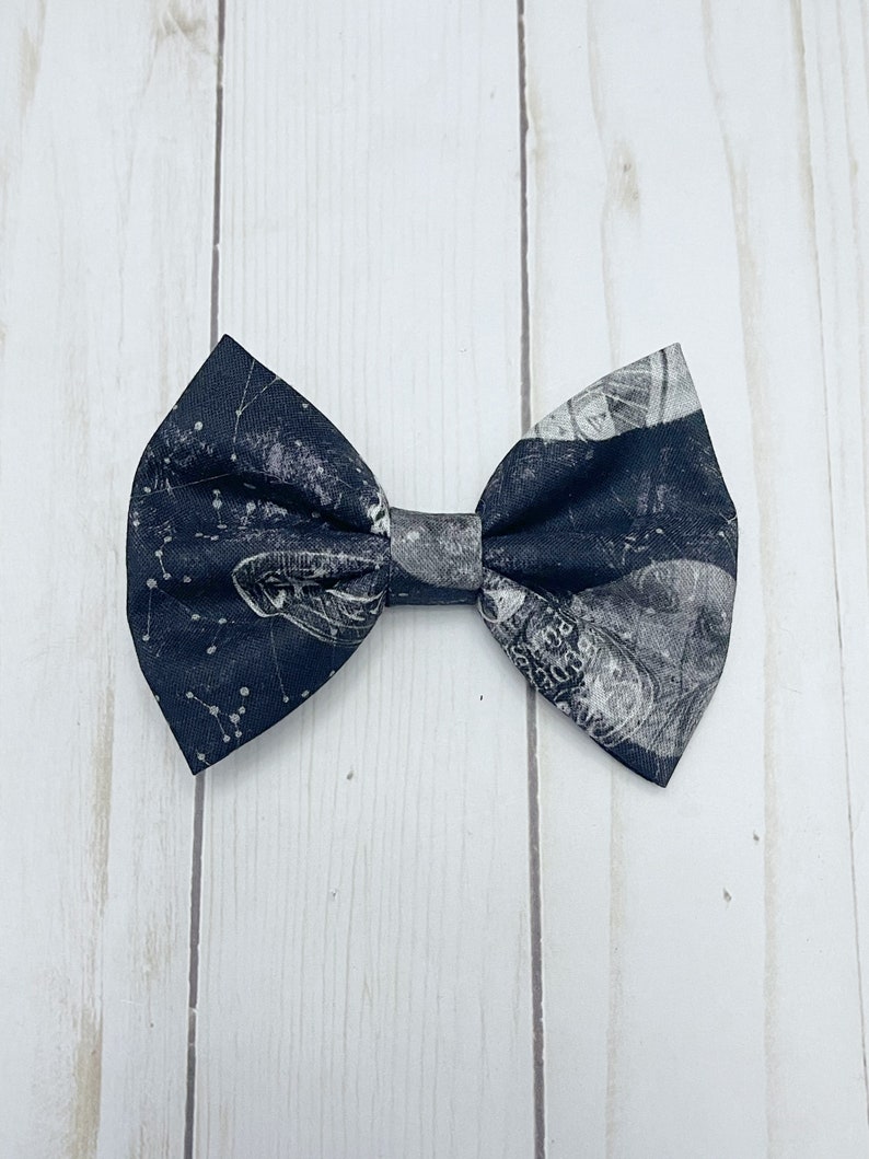 May include: A fabric bow tie featuring a dark navy blue background with a white celestial print of constellations, stars, and planetary or lunar imagery. The bow tie has a classic pleated center knot and pointed wing edges on both sides.