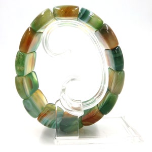May include: A green, brown, and white agate stone bracelet with rectangular beads. The bracelet is displayed on a clear plastic stand.