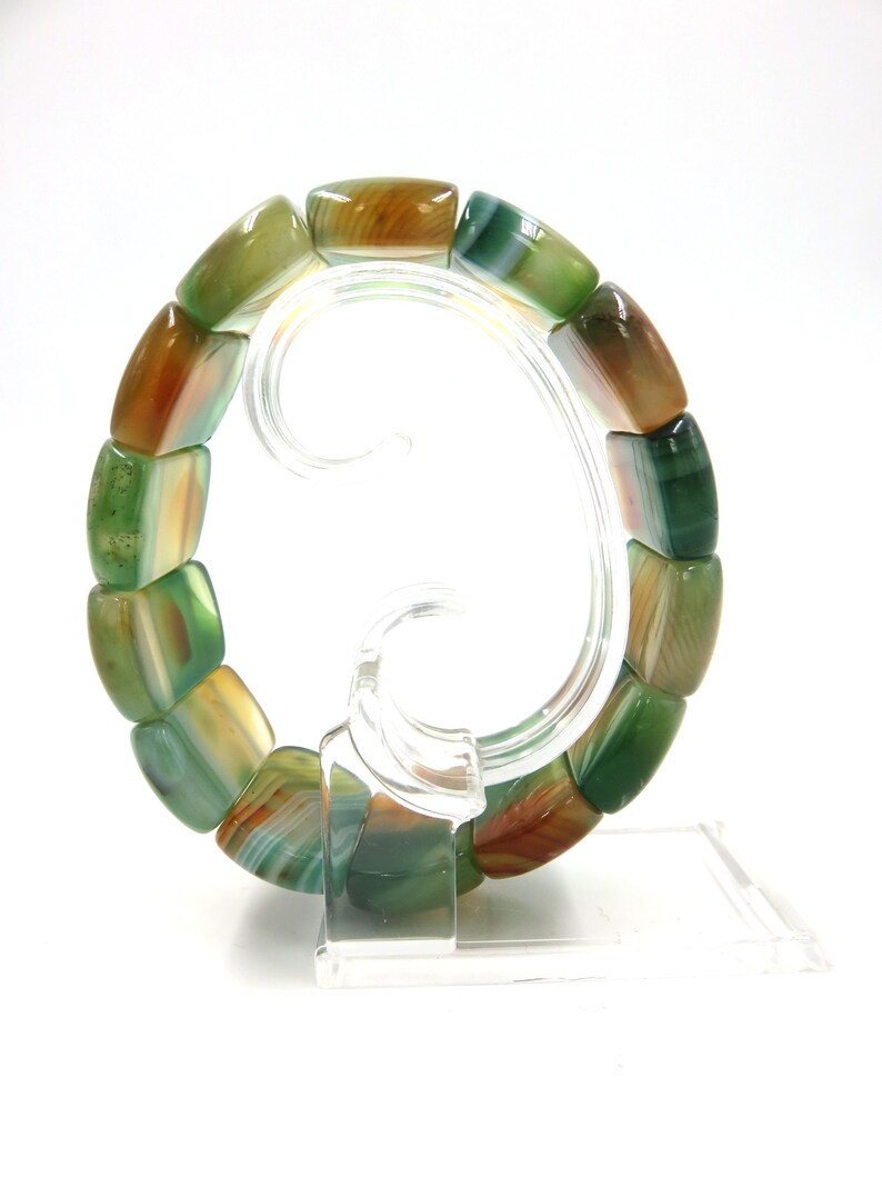 May include: A green, brown, and white agate stone bracelet with rectangular beads. The bracelet is displayed on a clear plastic stand.