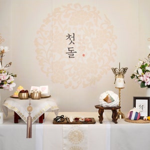 May include: A traditional Korean wedding ceremony setup with a white table cloth, two white vases with pink and white flowers, and a variety of traditional Korean wedding decorations. The text "첫돌" is written in black on a white background.