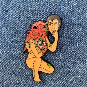 May include: A red and green enamel pin depicting a person with a creature growing out of their body. The creature has large eyes and is dripping green slime. The person is holding a donut and has a thoughtful expression.