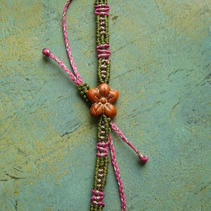 May include: A macrame bracelet featuring olive green cord woven with metallic pink thread. The centre is adorned with a five-petaled orange flower bead. The bracelet includes adjustable pink and metallic silver braided cord ties with knotted ends.