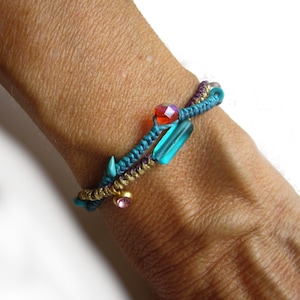 Könnte beinhalten: Ein mehrsträngiges Armband mit einer geflochtenen blaugrünen Kordel und einer goldfarbenen Metallkette. Das Design umfasst eine facettierte orangefarbene Perle, eine glatte rechteckige blaugrüne Perle und einen kleinen runden rosa Anhänger.