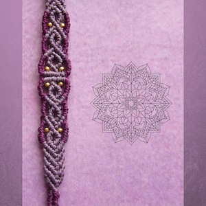 Könnte beinhalten: Ein handgefertigtes Makramee-Armband in Lila- und Grautönen, akzentuiert mit kleinen goldenen Perlen. Das Armband wird auf einem lavendelfarbenen Hintergrund neben einem schwarz-weißen Mandala-Design präsentiert.