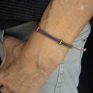 Könnte beinhalten: Ein handgefertigtes Armband mit einem gewebten Design in Lila-, Schwarz- und Olivgrüntönen. Das Armband hat goldfarbene Metallakzente und eine verstellbare Kordel in Beige. Das Armband wird am Handgelenk getragen.