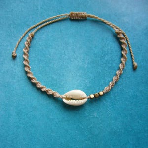 May include: A beige macrame cord bracelet featuring a single white cowrie shell and three small metallic gold-toned beads. The bracelet is constructed with a spiral knot pattern and includes an adjustable sliding knot closure at the top.