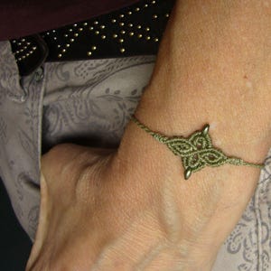 Könnte beinhalten: Ein handgefertigtes olivgrünes Makramee-Armband. Das Armband hat ein zentrales vierblättriges Kleeblatt-Design mit kleinen Metallperlen. Das Armband wird am Handgelenk getragen, mit einem Blick auf einen schwarzen, mit Nieten besetzten Gürtel und gemusterte Kleidung im Hintergrund.