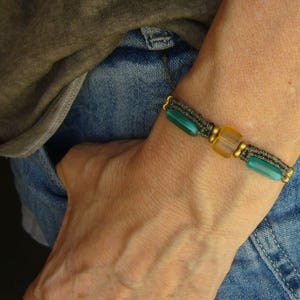 May include: A handmade bracelet featuring a dark gray woven band with teal and yellow beads. The bracelet is accented with gold-colored metal beads. The person is wearing a gray shirt and blue jeans.