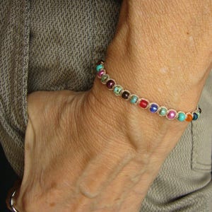 Può includere: Un braccialetto caratterizzato da una serie di piccole perline multicolori nelle tonalità del rosso, blu, verde e rosa, infilate su un cordino color marrone chiaro. Le perline sono disposte in un'unica fila, formando un semplice cerchio.