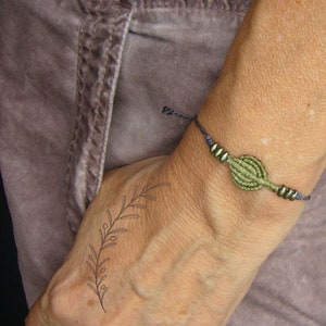May include: A macrame bracelet with a central olive green woven design and small metallic beads. The bracelet is on a dark blue cord. A black line drawing of a plant is on the wrist.