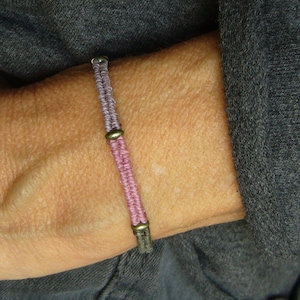 May include: A thin, woven cord bracelet featuring alternating sections of light purple, pink, and grey thread. The bracelet is accented with small, round, metallic bronze-toned beads placed between the different colored segments of the woven cord.