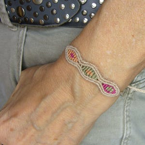 May include: A handmade macrame bracelet in beige with pink, orange, and green detailing. The bracelet is worn on a wrist and features a series of connected oval patterns. The bracelet has a unique, bohemian style and is a piece of handcrafted jewellery.