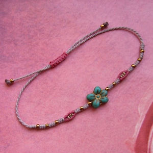May include: A delicate bracelet featuring a central turquoise-colored flower bead, accented with small metallic gold beads and pink thread details. The band is constructed from light gray cord with an adjustable sliding knot closure mechanism.