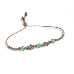 Può includere: Un braccialetto macramè fatto a mano con un cordoncino marrone chiaro, impreziosito da tre perline di vetro azzurro chiaro e piccole perline color bronzo. Il braccialetto regolabile è presentato su sfondo bianco. Questo braccialetto in stile bohémien è perfetto per l'uso quotidiano.