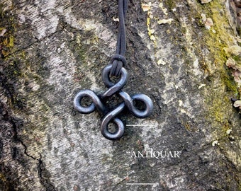 May include: A black metal pendant with a Celtic knot design hangs from a black cord. The pendant is on a tree trunk background.