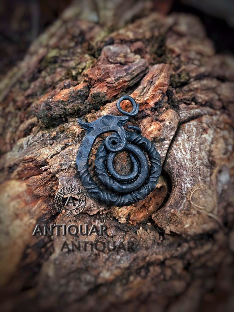 Puede incluir: Un colgante de metal negro con un dise&ntilde;o estilizado de serpiente o drag&oacute;n. El colgante est&aacute; sobre una superficie de madera.