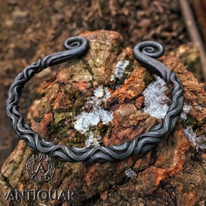 Puede incluir: Un brazalete de metal negro con un diseño retorcido en forma de onda y extremos curvados. El brazalete está apoyado en un trozo de corteza de árbol. La palabra "ANTIQUAR" es visible en la esquina inferior izquierda de la imagen.