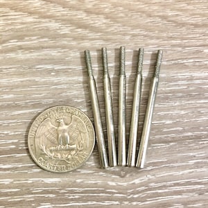 Może przedstawiać: Pięć srebrnych wierteł z teksturowanymi końcówkami ułożonych na drewnianej powierzchni obok ćwierćdolara amerykańskiego. Wiertła są cylindryczne. Moneta przedstawia orła.