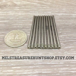 May include: A close-up of a set of 12 silver diamond drill bits, each about 2 inches long, arranged in a row on a wooden surface. A US quarter is included for size comparison.