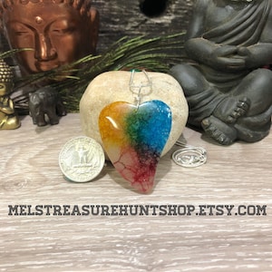 May include: A rainbow agate heart pendant on a silver chain. The pendant is about the size of a UK 20 pence coin. The pendant is on a wooden surface.