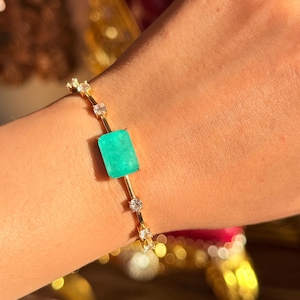 May include: A gold bracelet featuring a large, rectangular, bright green gemstone at its center. The bracelet is adorned with small, clear gemstones and is displayed on a person's wrist.