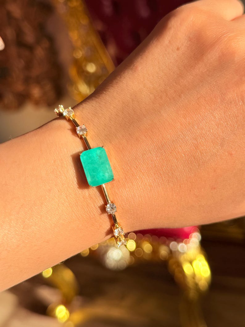 May include: A gold bracelet featuring a large, rectangular, bright green gemstone at its center. The bracelet is adorned with small, clear gemstones and is displayed on a person's wrist.