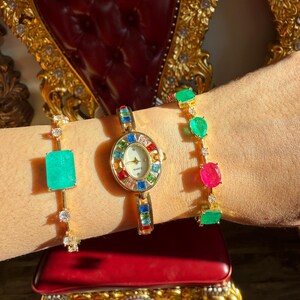 May include: A gold-toned watch and two bracelets adorned with colorful gemstones. The watch features a round face with a rainbow-colored bezel. One bracelet has a large emerald-colored stone, and the other has green and pink stones.