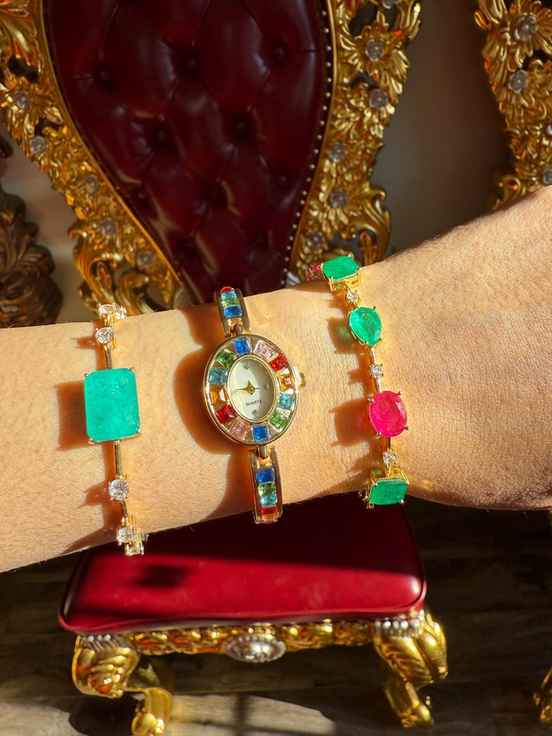 May include: A gold-toned watch and two bracelets adorned with colorful gemstones. The watch features a round face with a rainbow-colored bezel. One bracelet has a large emerald-colored stone, and the other has green and pink stones.