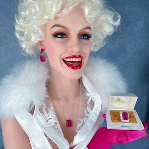 May include: A white blouse with a lace overlay and a pink bow. The blouse is worn by a mannequin with blonde curly hair and red lipstick. The mannequin is holding a small white box with a red jewel ring inside. The box has the text "Gemesis" on it.