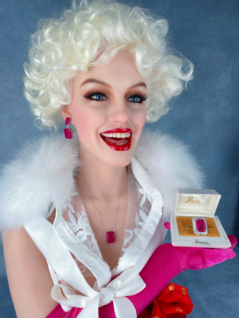 May include: A white blouse with a lace overlay and a pink bow. The blouse is worn by a mannequin with blonde curly hair and red lipstick. The mannequin is holding a small white box with a red jewel ring inside. The box has the text "Gemesis" on it.