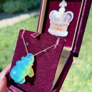 Puede incluir: Un collar de cadena dorada con un colgante de elefante iridiscente azul y amarillo. El collar está en una caja de terciopelo rojo con una corona en la tapa.