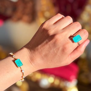 May include: A gold bracelet and ring set. The bracelet features a rectangular, light blue gemstone and small, round, clear stones. The ring has a matching rectangular light blue gemstone. The jewelry is displayed on a person's hand.