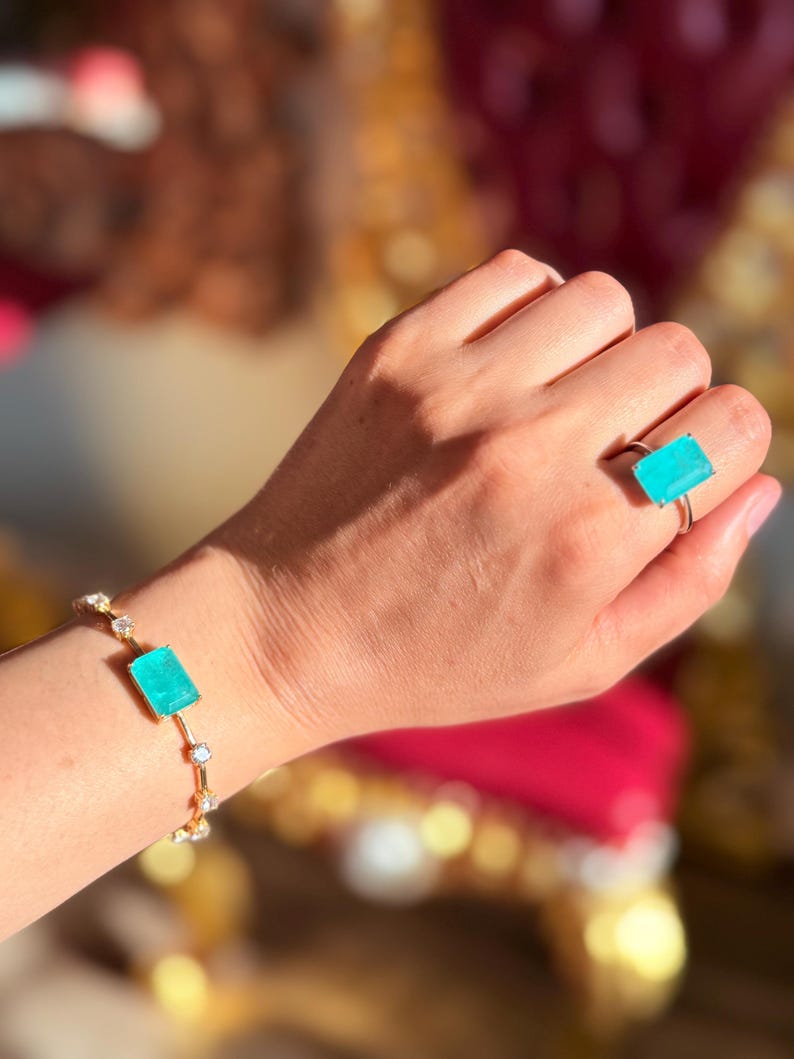May include: A gold bracelet and ring set. The bracelet features a rectangular, light blue gemstone and small, round, clear stones. The ring has a matching rectangular light blue gemstone. The jewelry is displayed on a person's hand.
