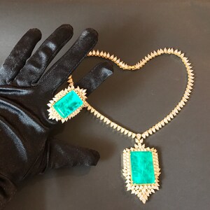 Op de afbeelding: Een gouden ketting met een grote hanger van smaragdgroene edelsteen en een bijpassende ring. De ketting is gemaakt van kleine, peervormige diamanten.