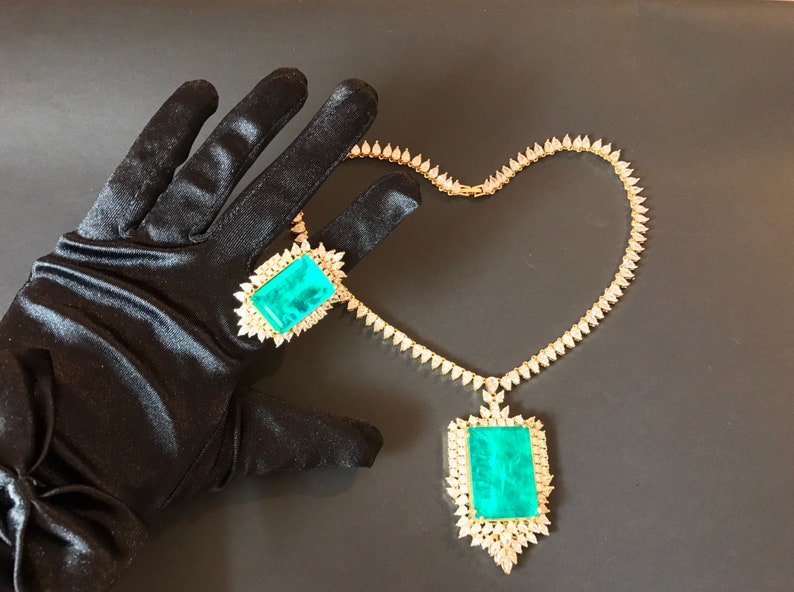 Op de afbeelding: Een gouden ketting met een grote hanger van smaragdgroene edelsteen en een bijpassende ring. De ketting is gemaakt van kleine, peervormige diamanten.