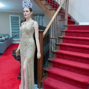 May include: A mannequin wearing a gold sequined gown with a high slit, a jeweled crown, and emerald earrings. The mannequin is standing on a red carpet, near a wooden staircase with a red runner.