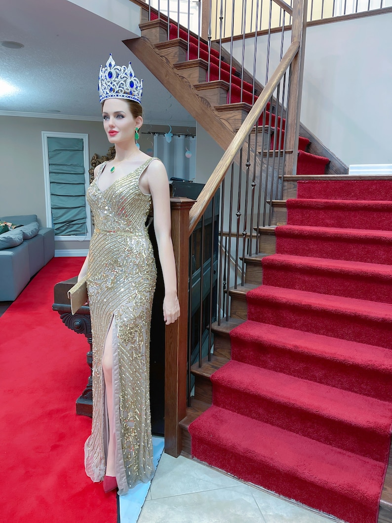 May include: A mannequin wearing a gold sequined gown with a high slit, a jeweled crown, and emerald earrings. The mannequin is standing on a red carpet, near a wooden staircase with a red runner.