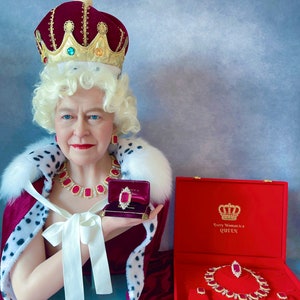 May include: A wax figure of a woman wearing a red velvet crown and robe, holding a red velvet jewelry box with a ruby ring inside. The box is open and the ring is visible. The text on the box reads "Every Woman is a Queen".