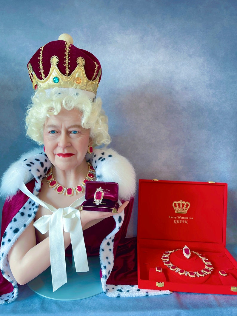 May include: A wax figure of a woman wearing a red velvet crown and robe, holding a red velvet jewelry box with a ruby ring inside. The box is open and the ring is visible. The text on the box reads "Every Woman is a Queen".