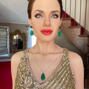 May include: A woman wearing a gold sequined dress, a gold necklace with a teardrop-shaped green pendant, and matching green gemstone earrings. The woman has red lipstick and is standing in front of a staircase.