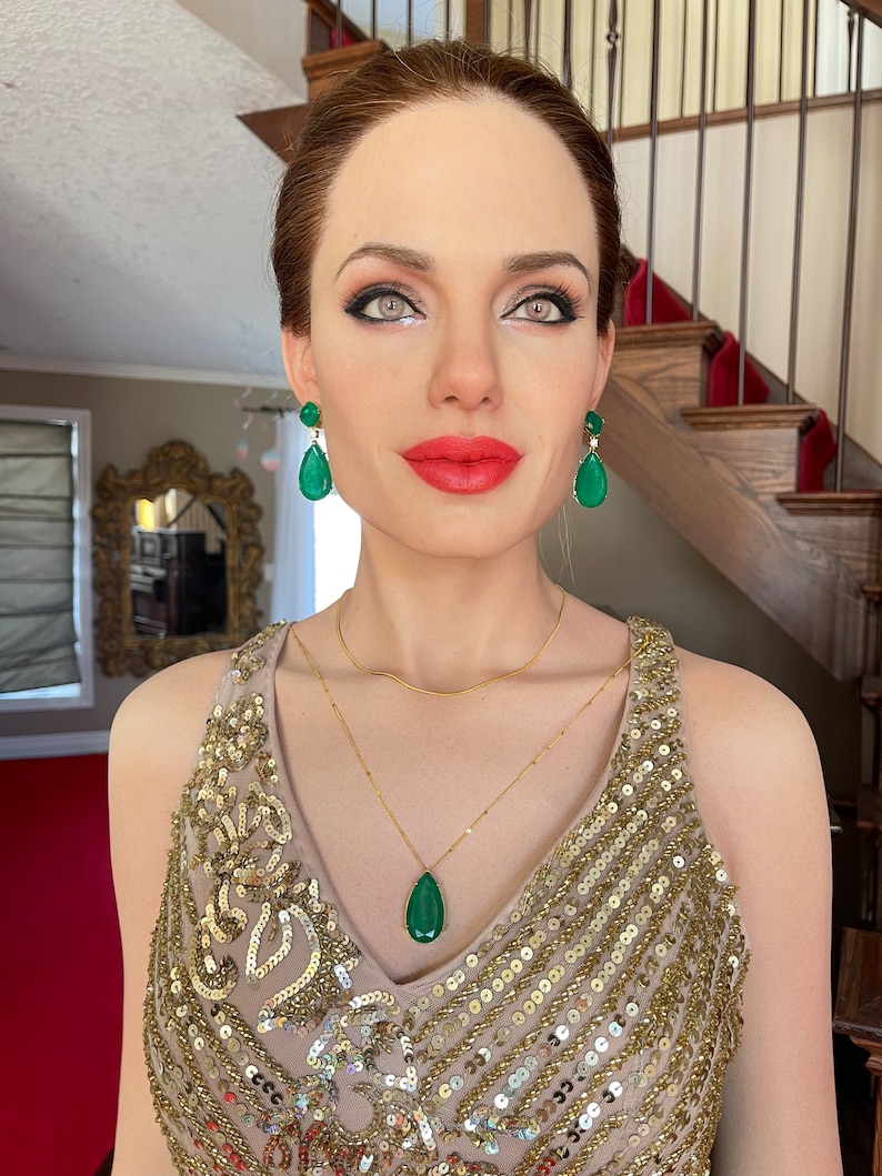 May include: A woman wearing a gold sequined dress, a gold necklace with a teardrop-shaped green pendant, and matching green gemstone earrings. The woman has red lipstick and is standing in front of a staircase.