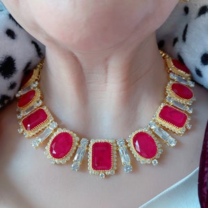 May include: A gold necklace with red and clear gemstones. The necklace features a row of rectangular red stones surrounded by smaller clear stones. The necklace is on a person's neck.