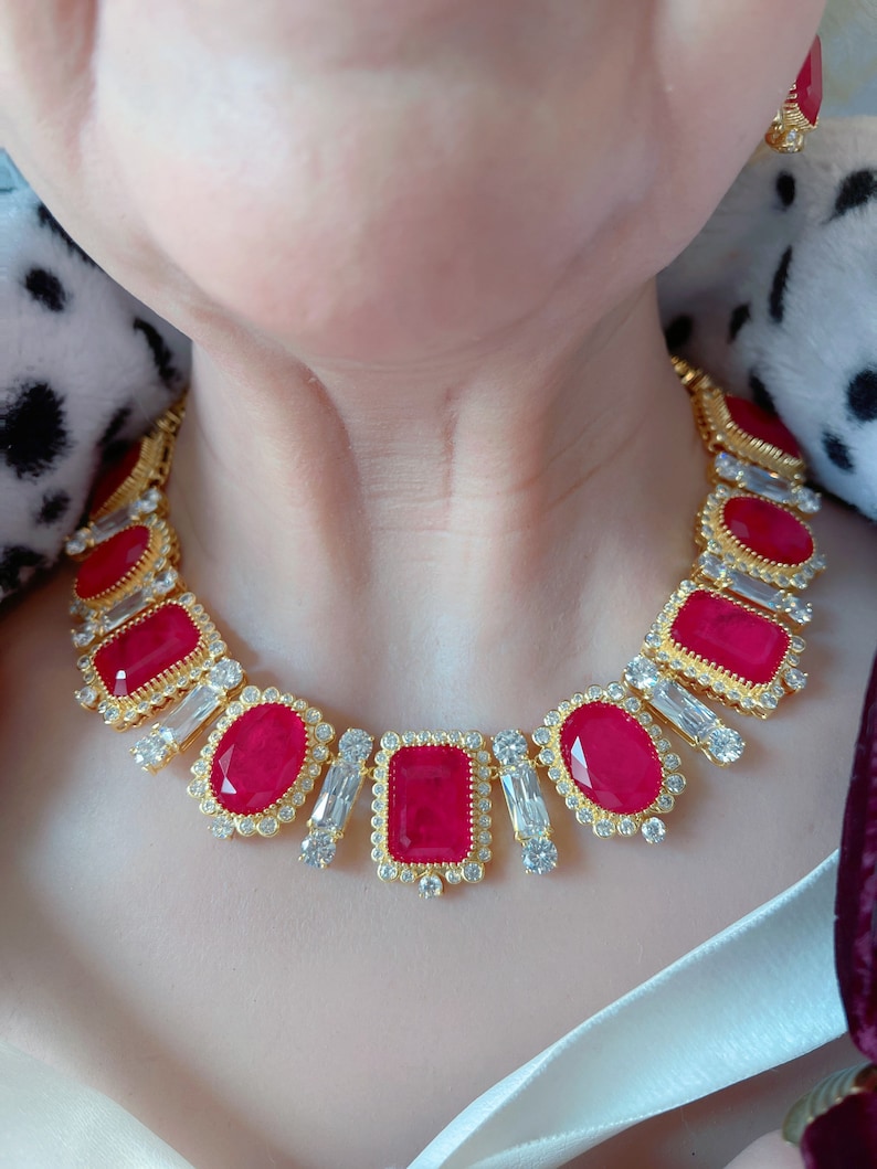 May include: A gold necklace with red and clear gemstones. The necklace features a row of rectangular red stones surrounded by smaller clear stones. The necklace is on a person's neck.