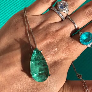 May include: A silver necklace with a large, teardrop-shaped, emerald-green gemstone pendant. Several rings with clear and blue gemstones adorn the hand. The jewelry is displayed against a turquoise background.