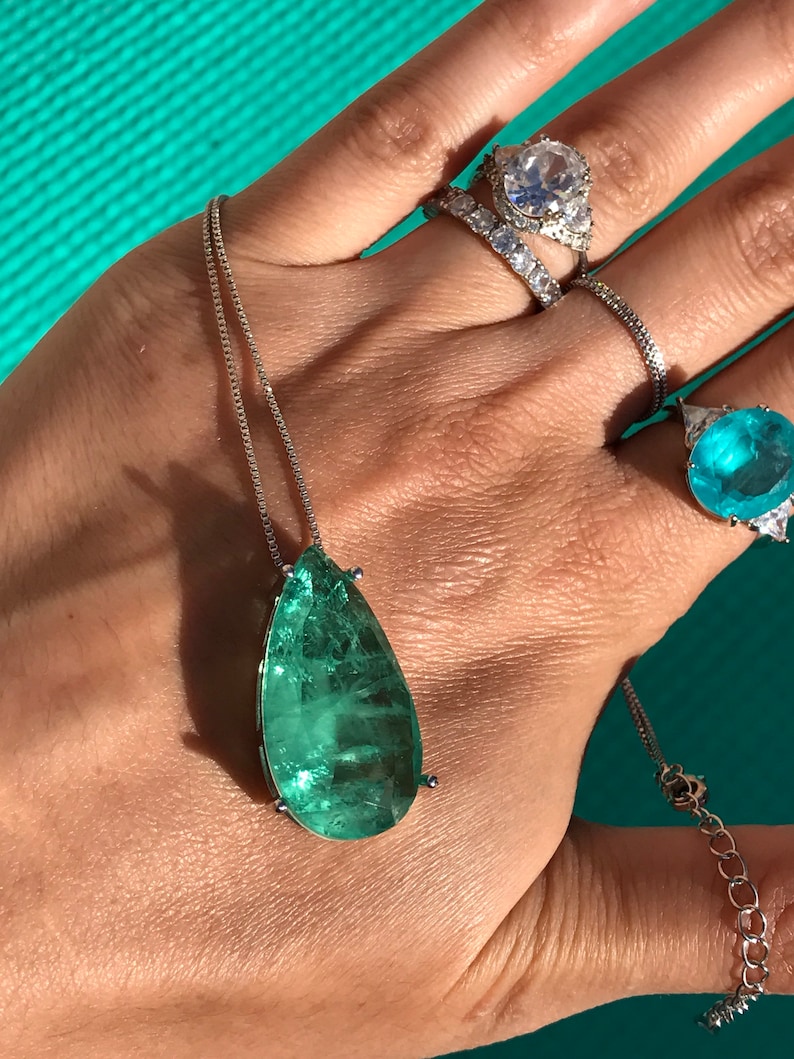 May include: A silver necklace with a large, teardrop-shaped, emerald-green gemstone pendant. Several rings with clear and blue gemstones adorn the hand. The jewelry is displayed against a turquoise background.