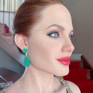 May include: A side profile of a person wearing emerald green drop earrings and a gold necklace. The person has red lipstick and dark eye makeup. The background includes a red carpet and a staircase.