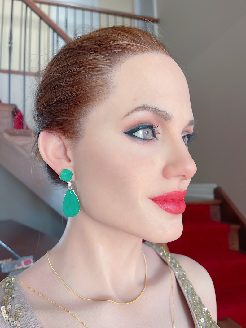 May include: A side profile of a person wearing emerald green drop earrings and a gold necklace. The person has red lipstick and dark eye makeup. The background includes a red carpet and a staircase.