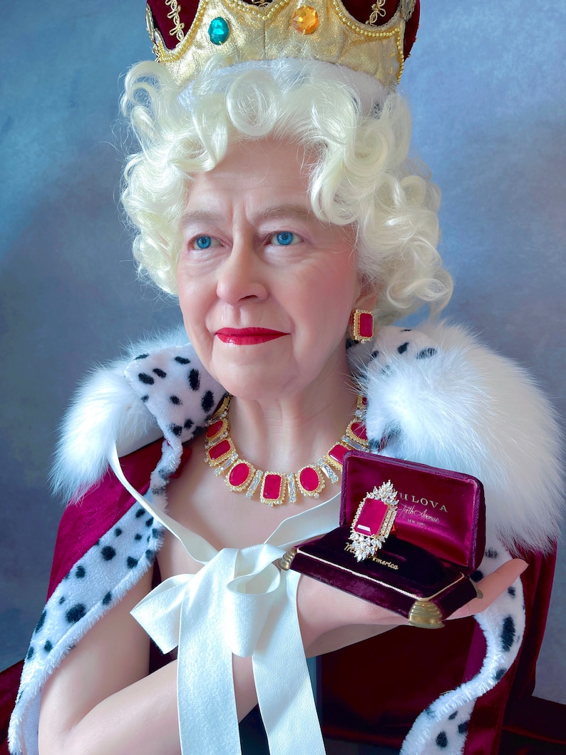 May include: A woman wearing a crown and a red and white fur-trimmed robe holds a red velvet box with a large ruby ring inside. The ring is surrounded by smaller diamonds.