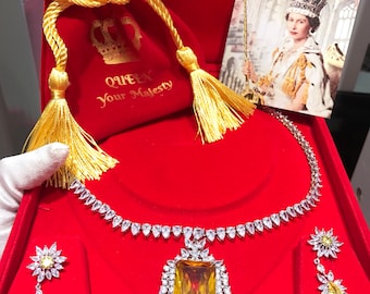 May include: A red velvet jewelry box with a gold trim and a gold crown design. The box contains a gold and white necklace with a large yellow gemstone pendant and matching earrings. The box also includes a small red velvet bag with a gold drawstring and a gold crown design. The bag has the text "Queen Your Majesty" printed on it. A small card with a picture of Queen Elizabeth II is also included in the box.